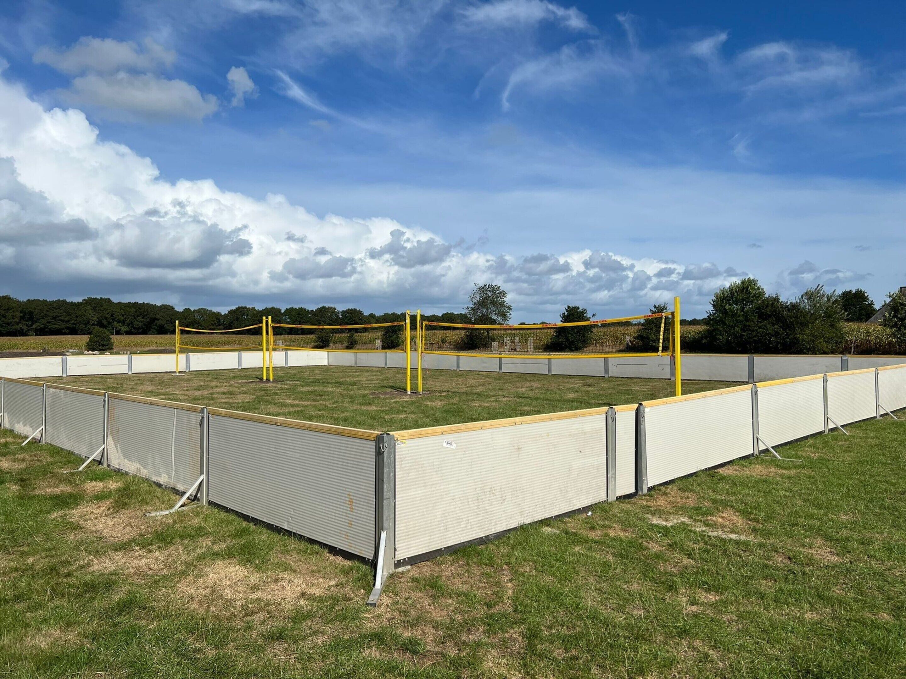 Un terrain de sport clôturé, équipé d’un filet de volley-ball, entouré d’un champ herbeux et d’arbres.