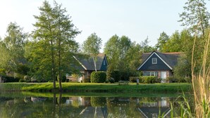 Cena tranquila à beira de um lago, com duas casas – uma de telhado escuro e outra com telhado mais claro – rodeadas por vegetação exuberante e um lago calmo que reflete as construções.