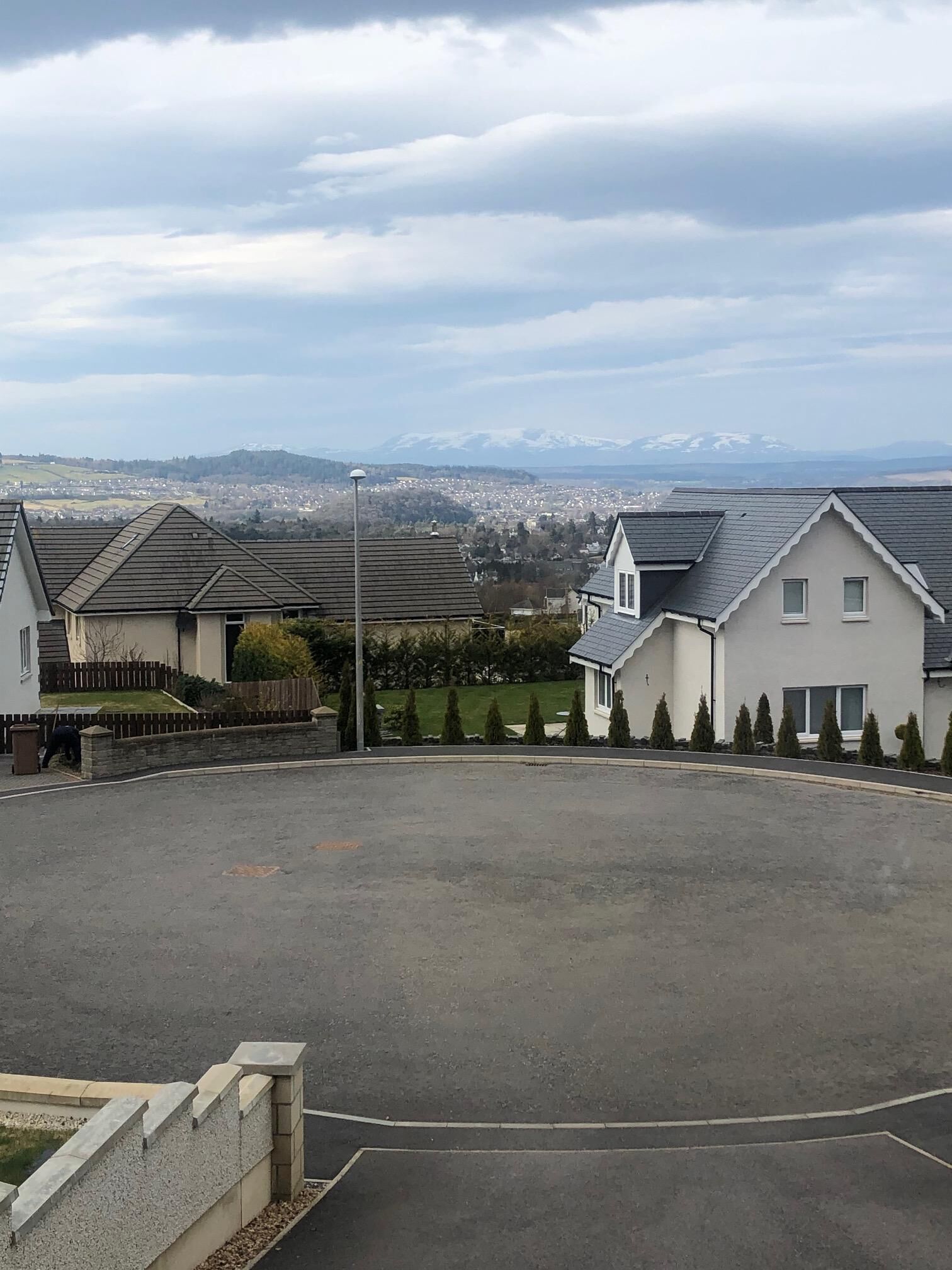 A residential area with houses, a paved driveway, and a distant view of a town and mountains.