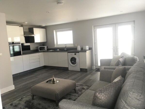 A modern kitchen with a washing machine, a microwave, and a sink. There is a grey sofa and a rug in the living area. - Appin Apartment (Inverness)