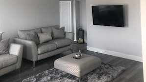 A modern living room with a grey sofa, a matching ottoman, a flat-screen TV mounted on the wall, and a patterned rug on the floor. - Appin Apartment (Inverness)
