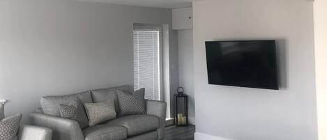 Una sala de estar moderna con un sofá gris, un reposapiés a juego, un televisor de pantalla plana montado en la pared y una alfombra estampada en el suelo.