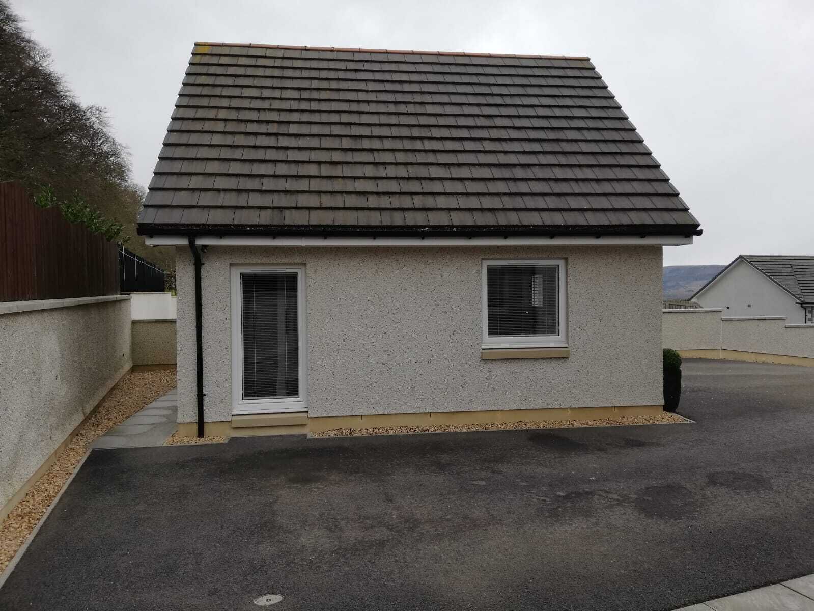 A small, single-story house with a dark shingle roof, two windows, and a concrete driveway.