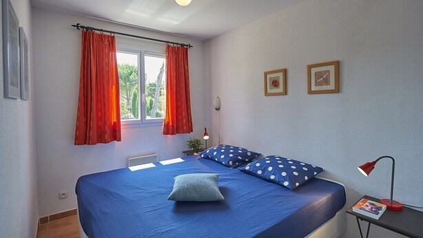 Une chambre à coucher avec un lit, une fenêtre avec des rideaux, une table de chevet avec une lampe, et deux tableaux encadrés accrochés au mur.