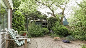 A garden with a wooden bench and a small table, surrounded by various plants and a brick pathway. - Cozy Holiday Home in Noordwijkerhout near Lake (Noordwijkerhout)