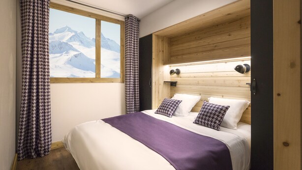 A bedroom with a wooden headboard, a bed with white and purple bedding, a window with a mountain view, and houndstooth curtains. - Alpine Stay w/ Spa + Views (Tignes)