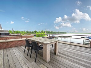 Una terraza de madera con una mesa y sillas, con vistas a una masa de agua tranquila con barcos y vegetación al fondo.