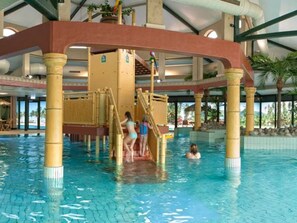Una piscina interior con un tobogán acuático en el centro, rodeada de pilares y una terraza cubierta.