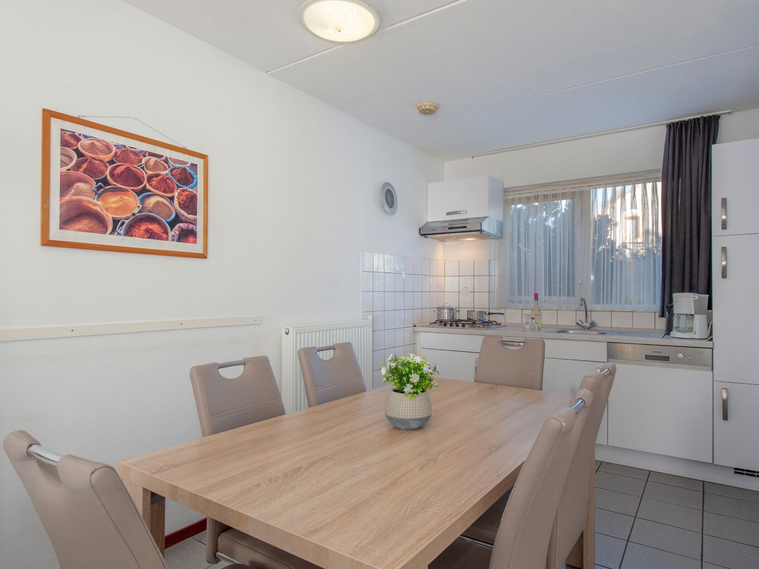Una cocina moderna con armarios blancos, estufa y fregadero. Se observa una mesa de comedor con sillas y una planta en maceta.