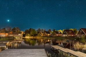 A lakeside view with wooden docks, a calm water surface reflecting the night sky, and houses with visible roofs and windows. - Lovely Tent with Kitchen & Bathroom Located near Pond (Bant)
