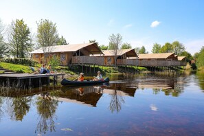 A riverside accommodation with wooden deck and canopies, a calm water body, and greenery.