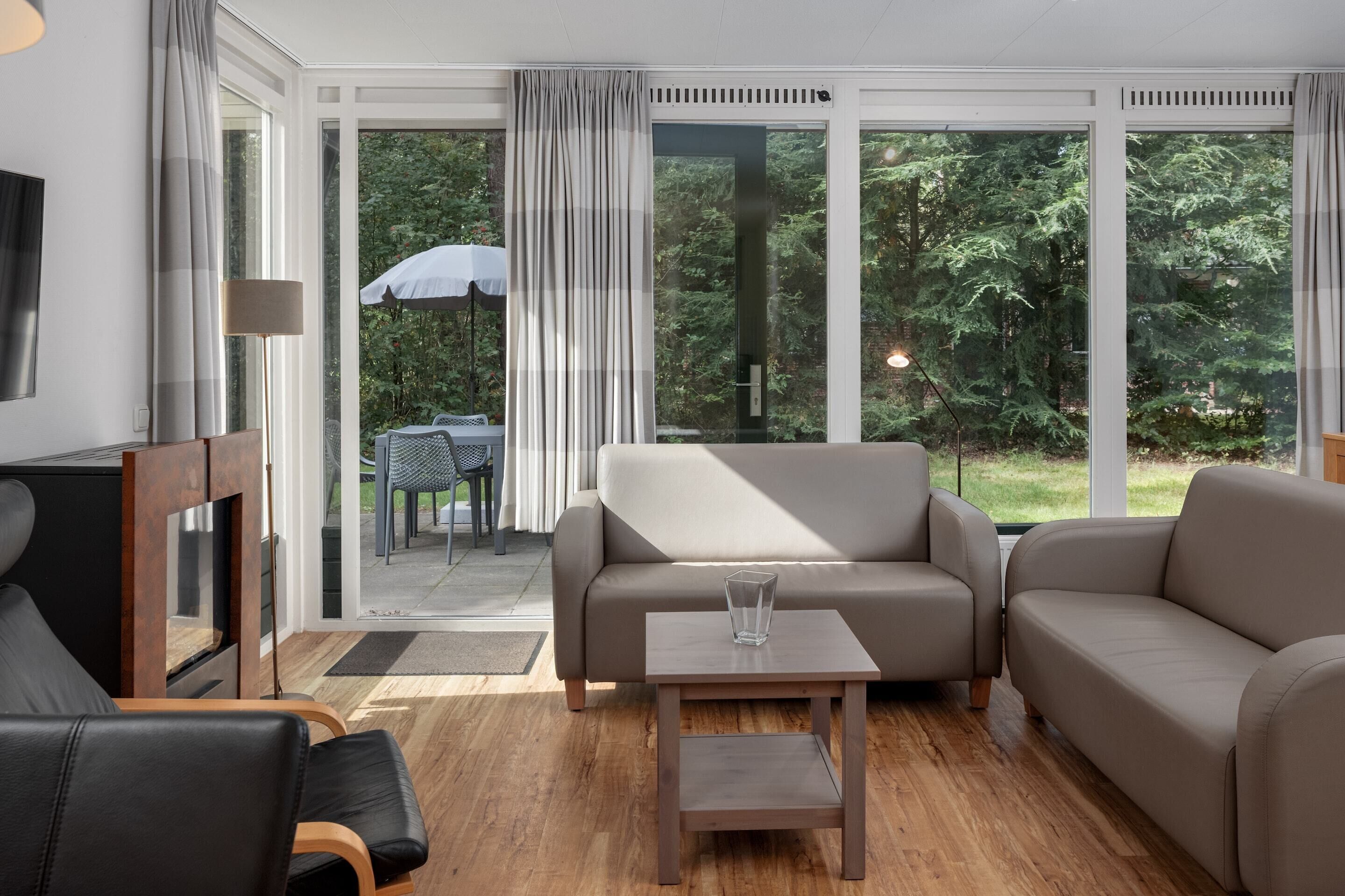 A modern living room with a sofa, a coffee table, and a TV. There are large windows with curtains, a view of greenery outside.