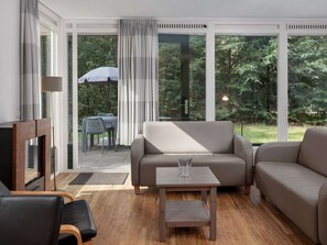 A modern living room with a sofa, a coffee table, and a TV. There is a large window with curtains, a dining table with chairs, and a garden view. - Chalet With Dishwasher Near Nature Reserve (Borger)
