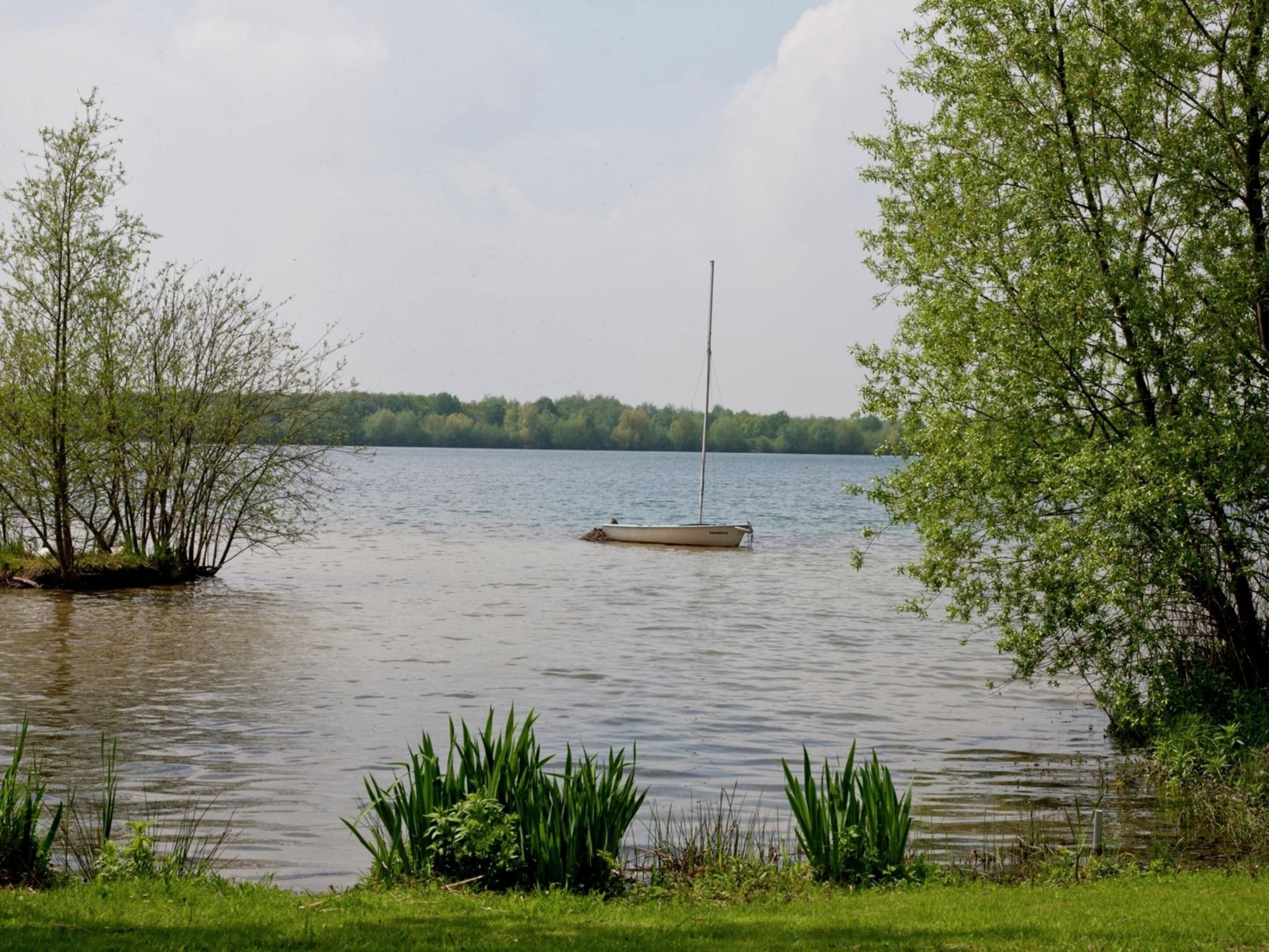 A calm lake with a small sailboat anchored near the shore, surrounded by lush greenery and trees.