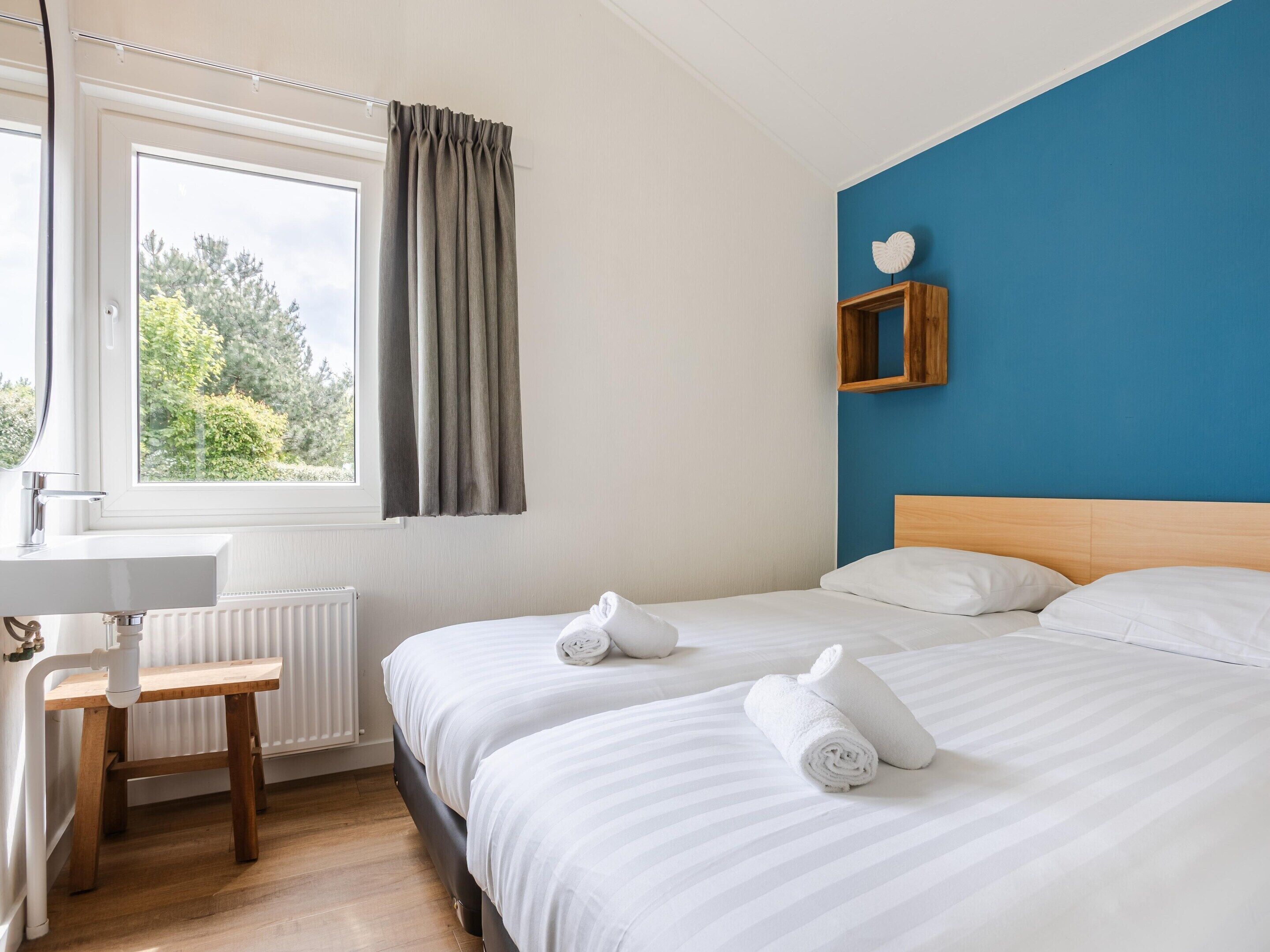 A hotel room with a bed, a small desk, a chair, a window with curtains, and a blue accent wall.