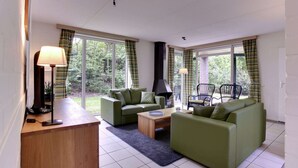 Ein modernes Wohnzimmer mit grünen Sofas, einem Holztisch und einer Lampe. Große Fenster mit Vorhängen bieten Blick auf Grünflächen außerhalb.