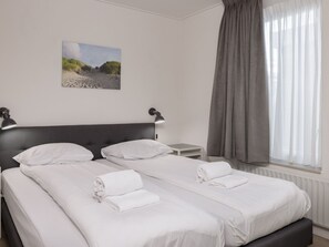 A double bed with white linens and two pillows, a bedside table with a lamp, a painting of a beach scene, and a window with grey curtains. - Bungalow in Netherlands With Private Sauna (Kamperland)