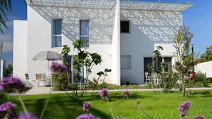 En modern vit byggnad med ett mönstrat solskydd, uteservering och en välskött gräsmatta med blommor.