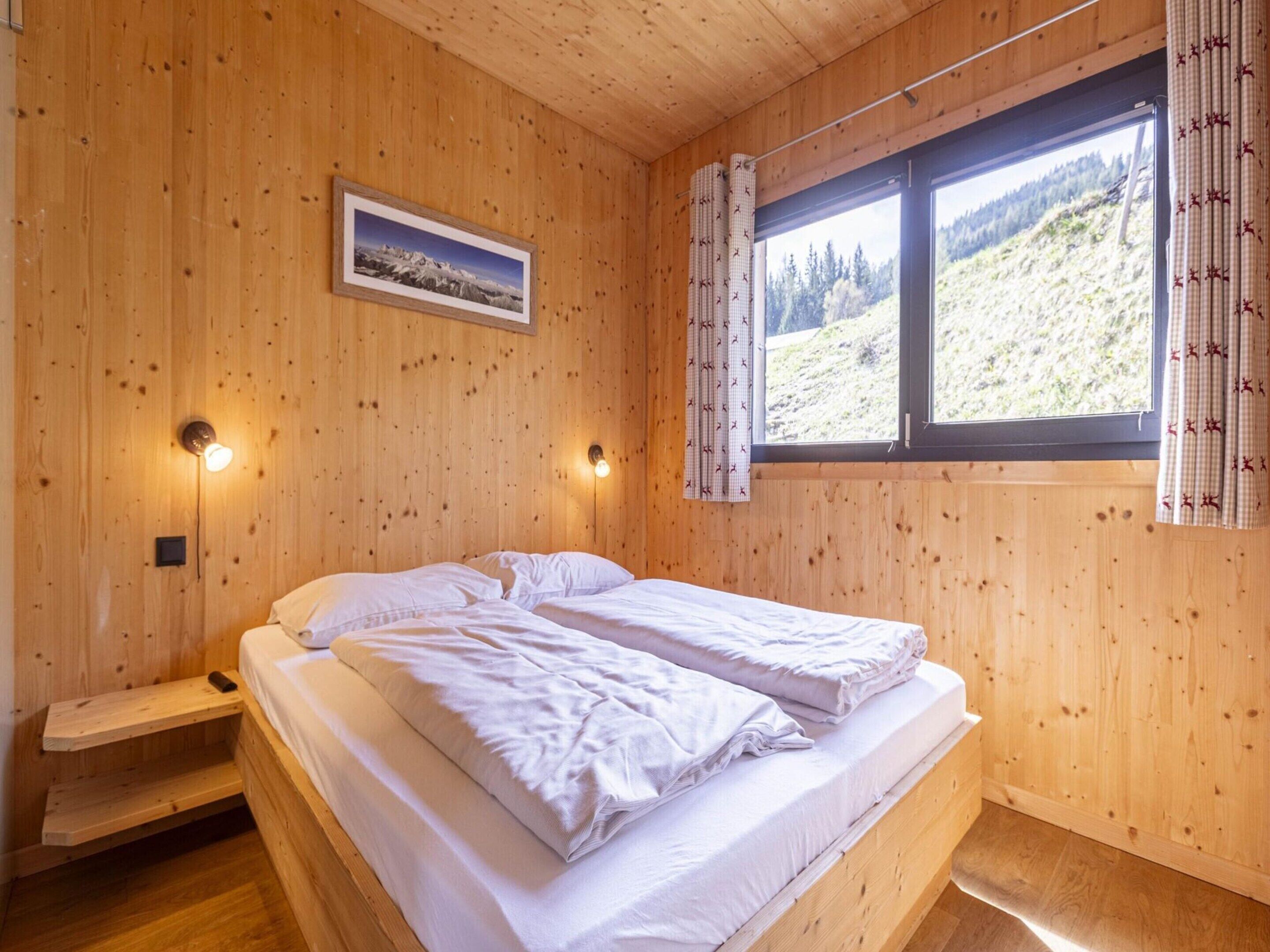 A wooden cabin bedroom with a bed, two wall-mounted lights, a window with curtains, and a framed picture on the wall.