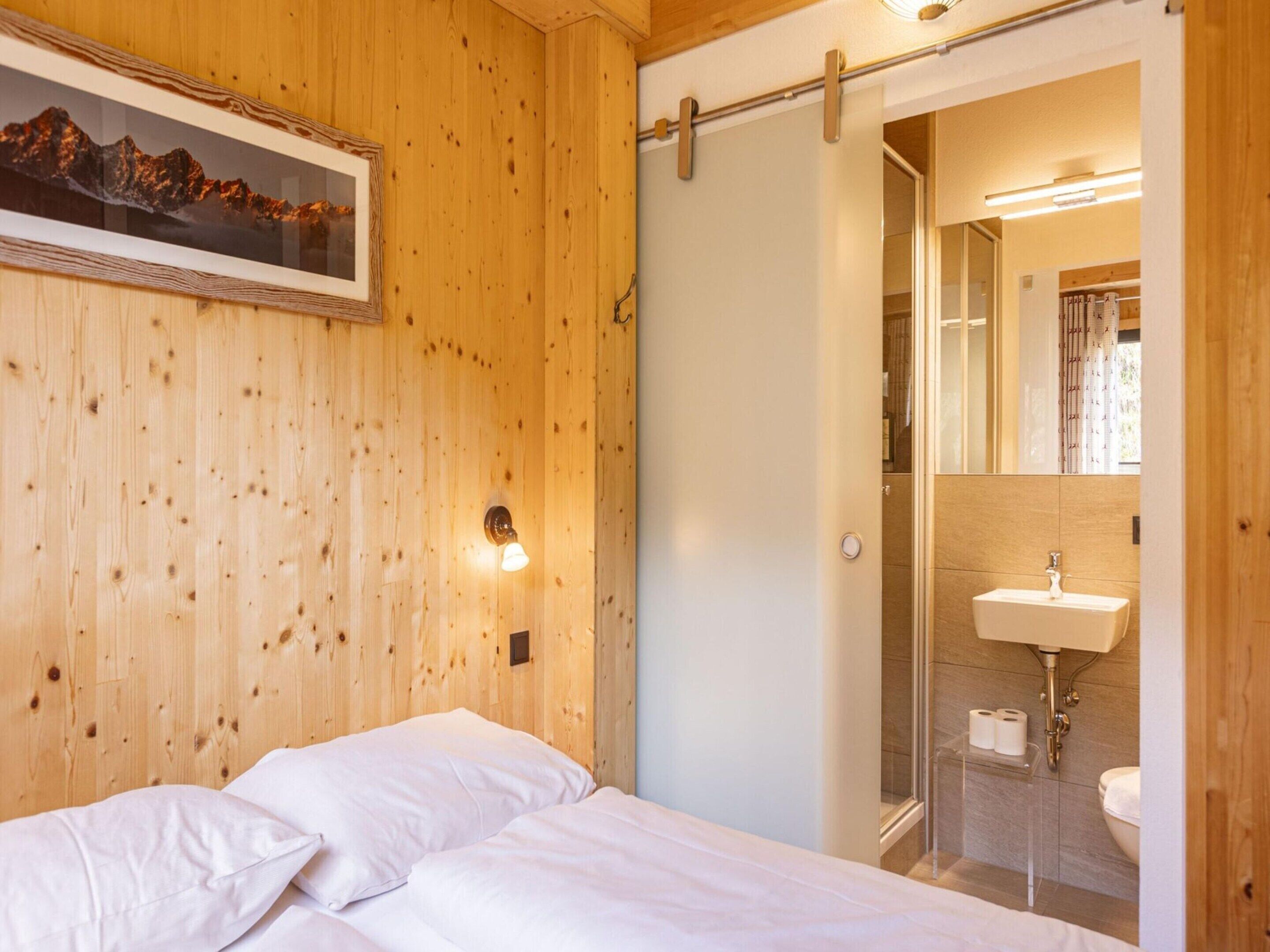 A wooden-paneled room with a bed, a framed picture, a wall-mounted light, and a bathroom visible through an open door.