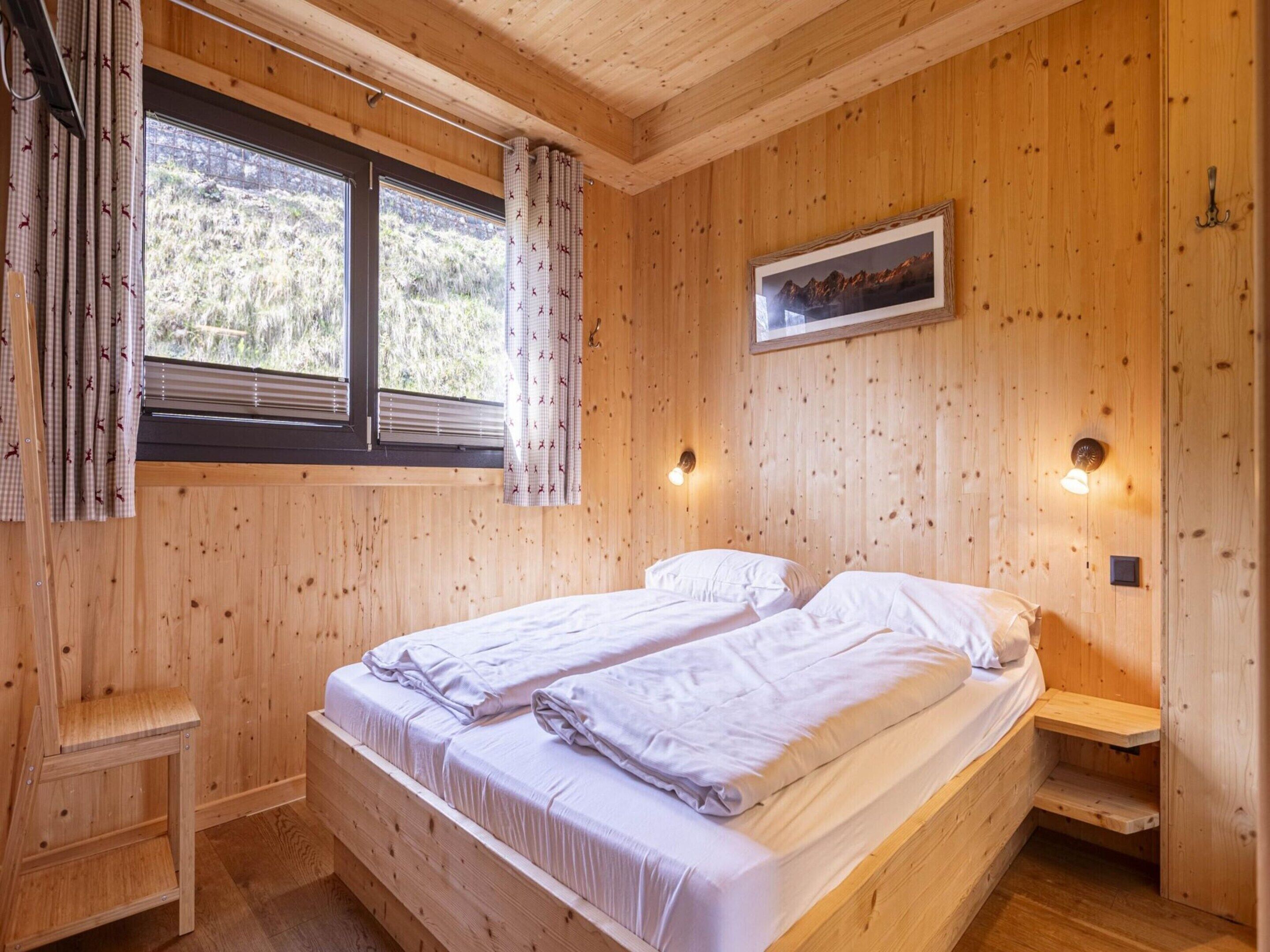 A wooden cabin room with two beds, a window with curtains, and a framed picture on the wall.