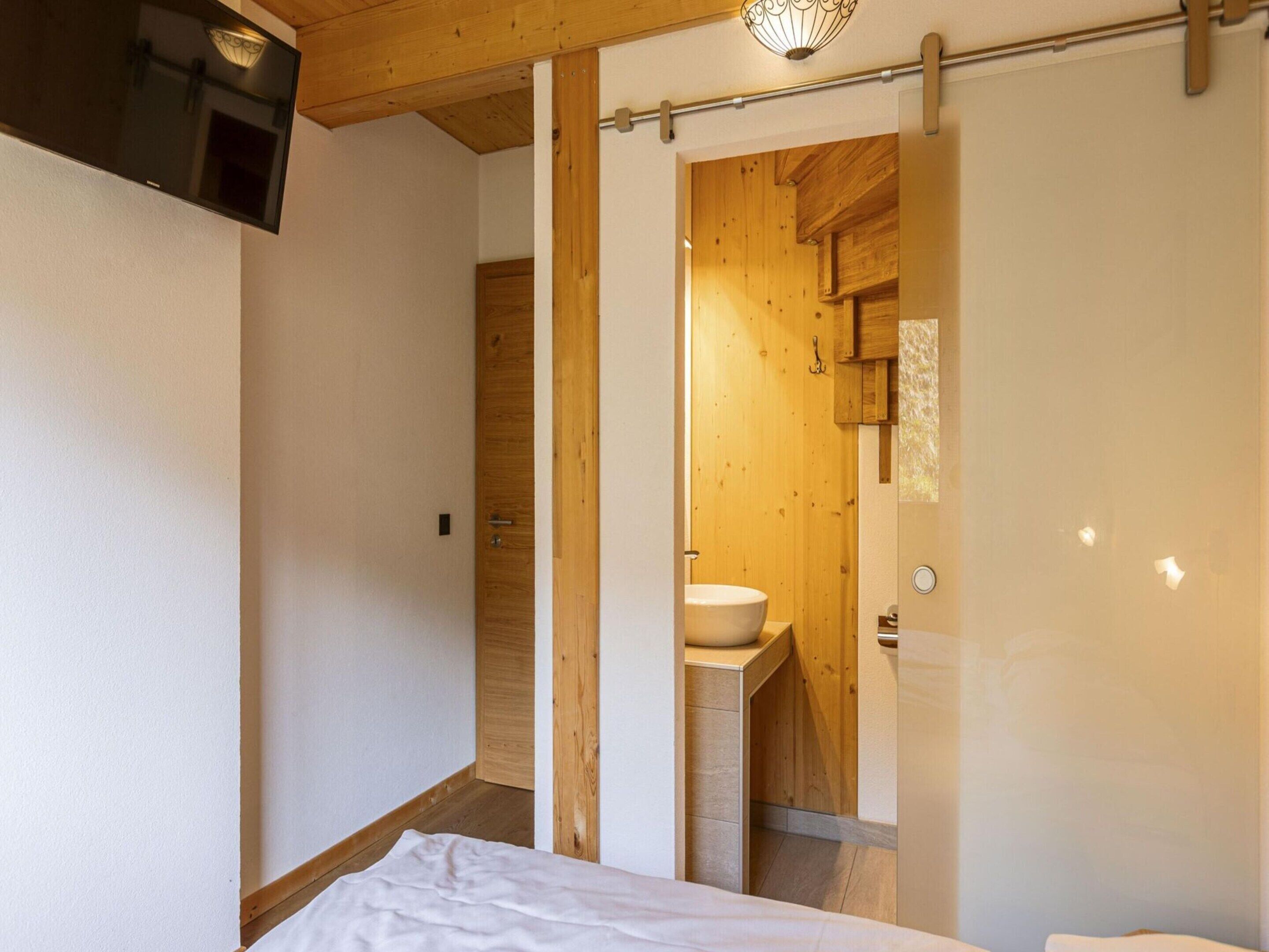 Une chambre à coucher comprenant un lit, un plafond en bois, un téléviseur fixé au mur et une porte coulissante donnant sur une salle de bain au décor intérieur en bois et dotée d’un lavabo.