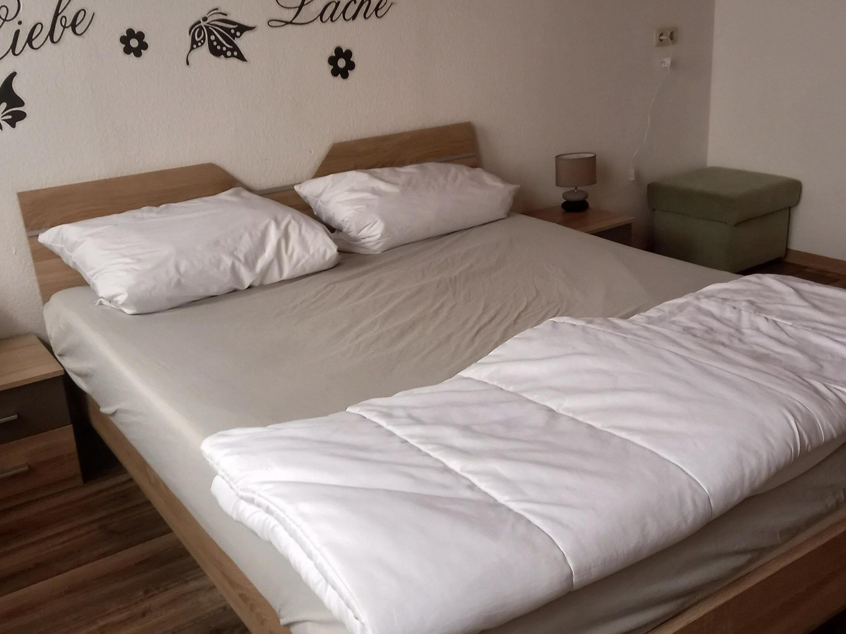 A single bed with white bedding, a wooden headboard, a bedside table with a lamp, and a green ottoman.