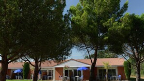 A building with a red-tiled roof, outdoor seating with blue umbrellas, and surrounding trees. - Family-friendly Apartment (Salleles-d'Aude)