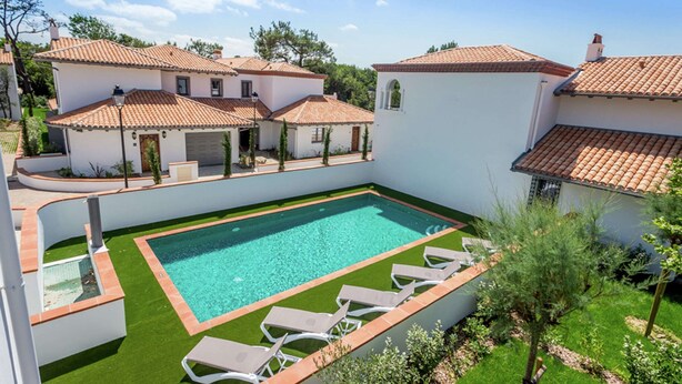 Une piscine entourée de chaises longues, un bâtiment blanc avec un toit en tuiles et un jardin.