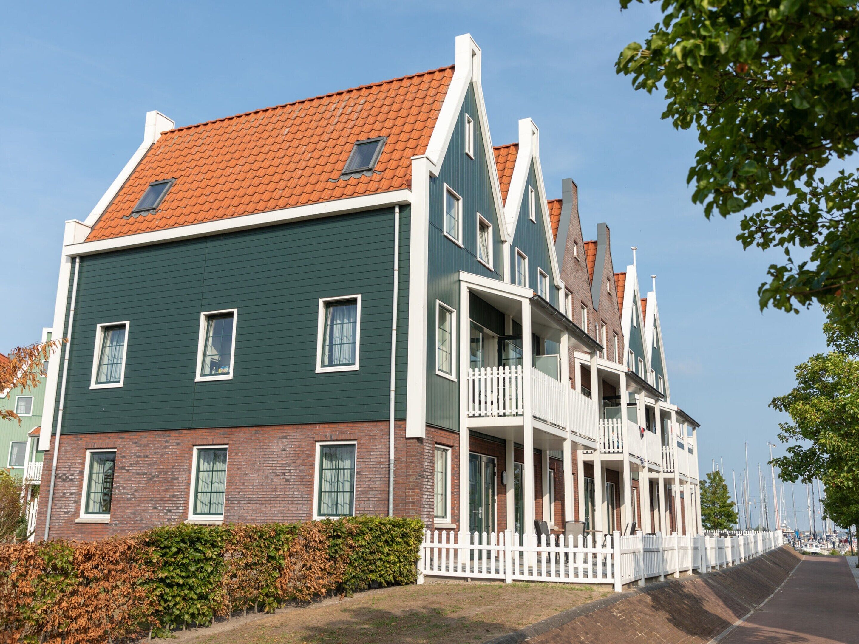 A multi-story building with a green facade and red-tiled roof, featuring balconies and a white picket fence.