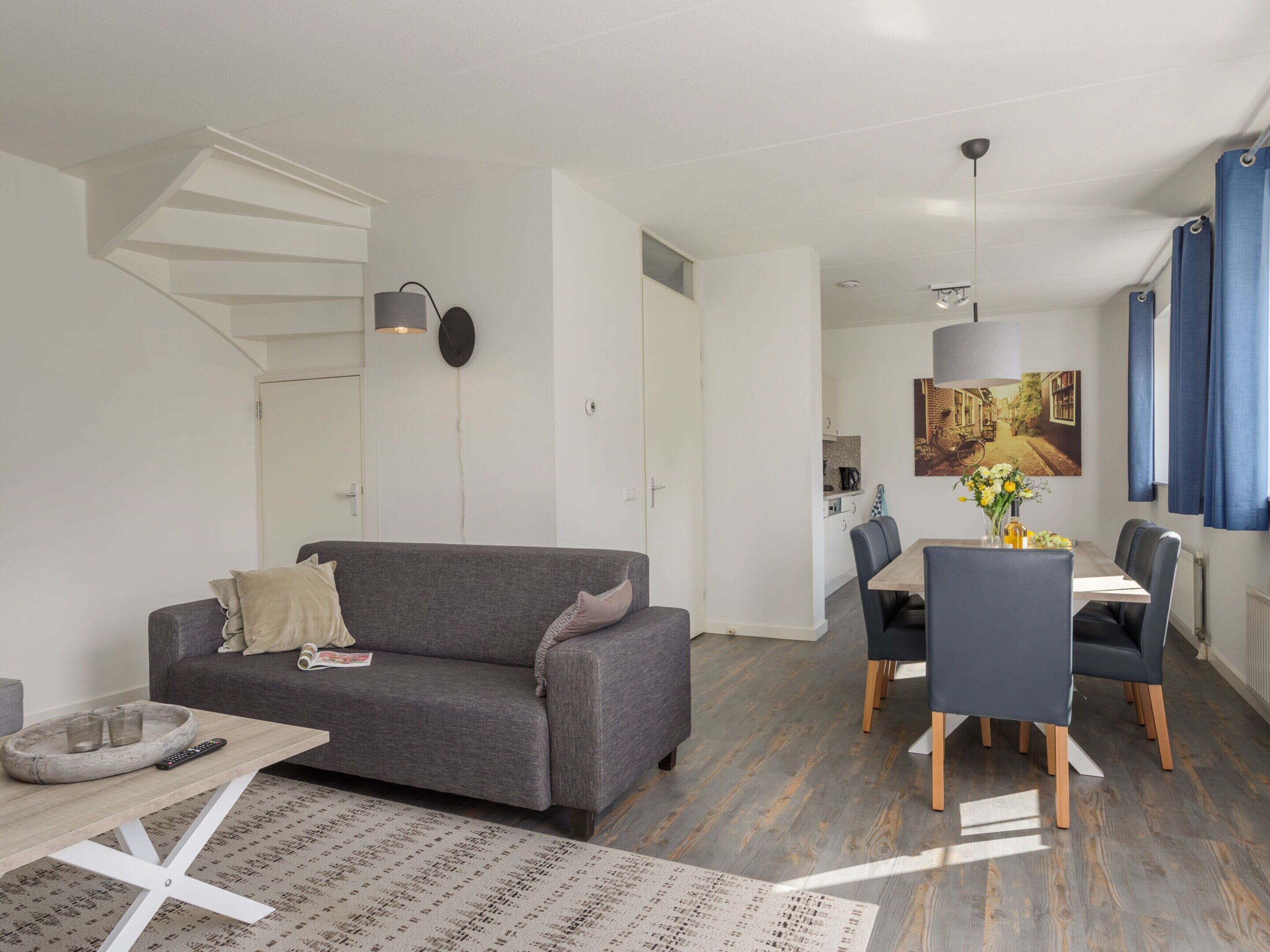 A modern living room with a grey sofa, a dining table with chairs, a kitchen in the background, and a staircase leading to another level.