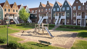 Un terrain de jeux avec des toboggans et un banc, entouré de maisons traditionnelles aux toits pentus et aux balustrades blanches.