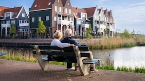 Un couple est assis sur un banc, près d’un canal, avec des maisons traditionnelles en arrière-plan.