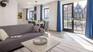 Un salon moderne avec un canapé gris, une table basse et un coin repas comprenant une table et des chaises. Une grande fenêtre ornée de rideaux bleus offre une vue sur un bâtiment à l’extérieur.
