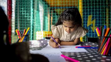 A child writing at a table with colorful pens and pencils, surrounded by a wire mesh and a yellow wall.