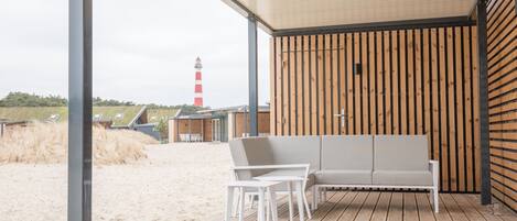 A covered outdoor seating area with a wooden floor, white chairs, and a sofa. A lighthouse is visible in the background.