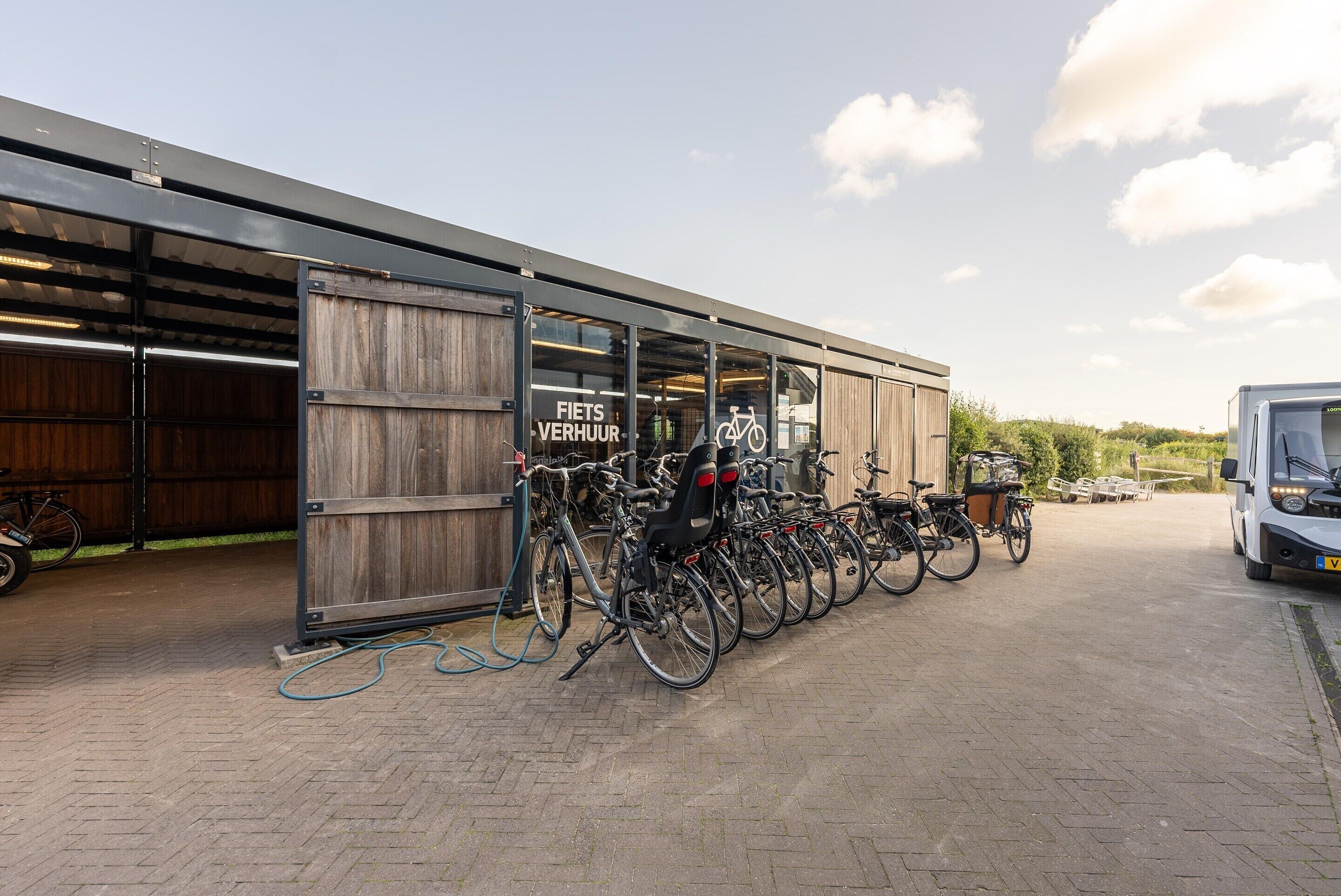 A bike rental station with multiple bicycles parked in front of a building with a sign reading "FIETS VERHUUR".