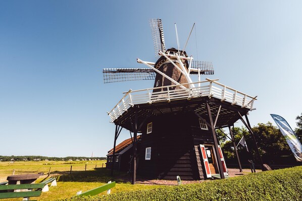Un moulin à vent traditionnel, doté d’une structure en bois et d’un toit en métal, entouré d’une zone herbeuse et surplombant un ciel dégagé.