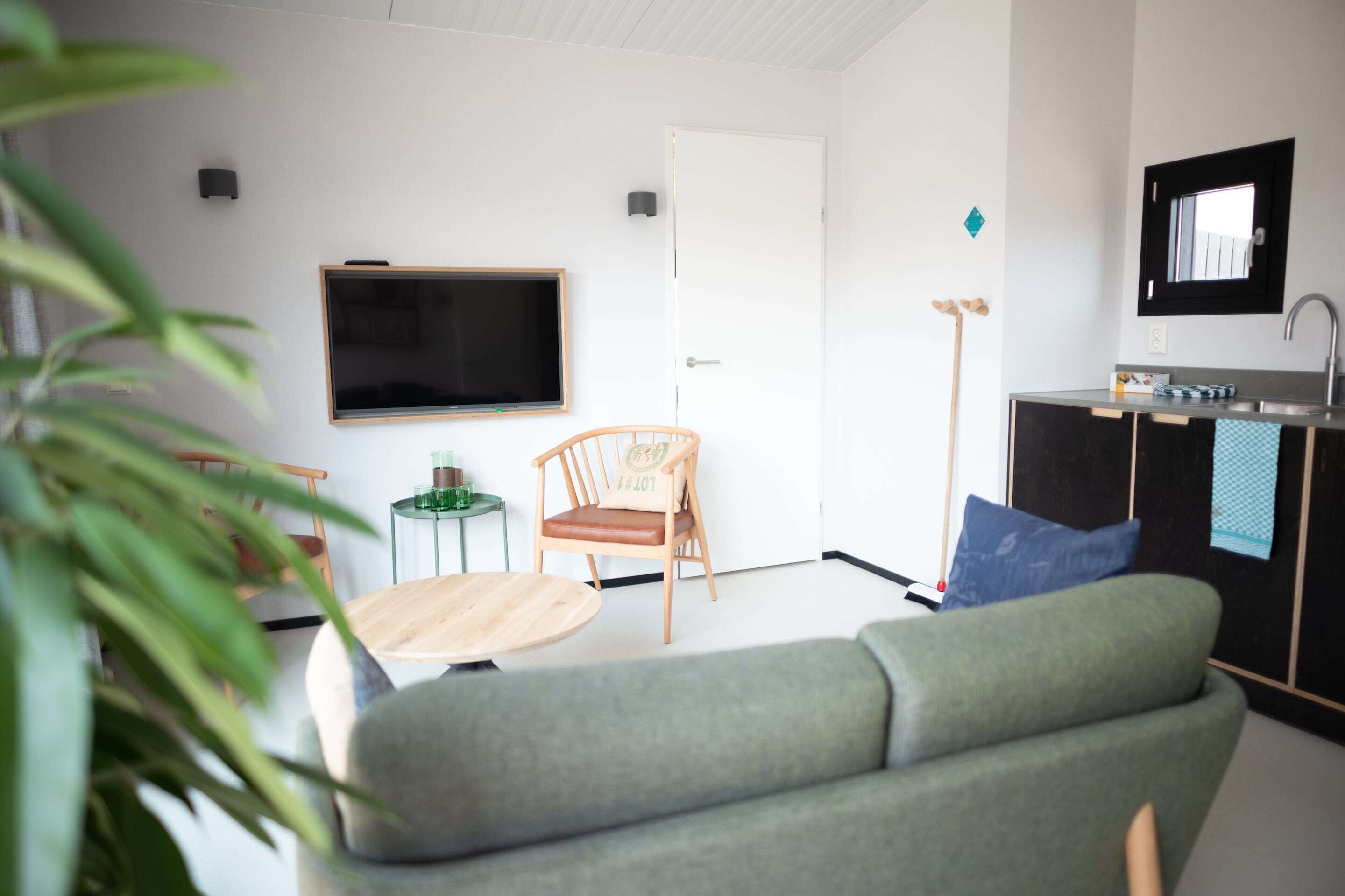 A modern living room with a grey sofa, a wooden chair, a small round table, a flat-screen TV, a kitchen area with a sink and countertop, and a door.