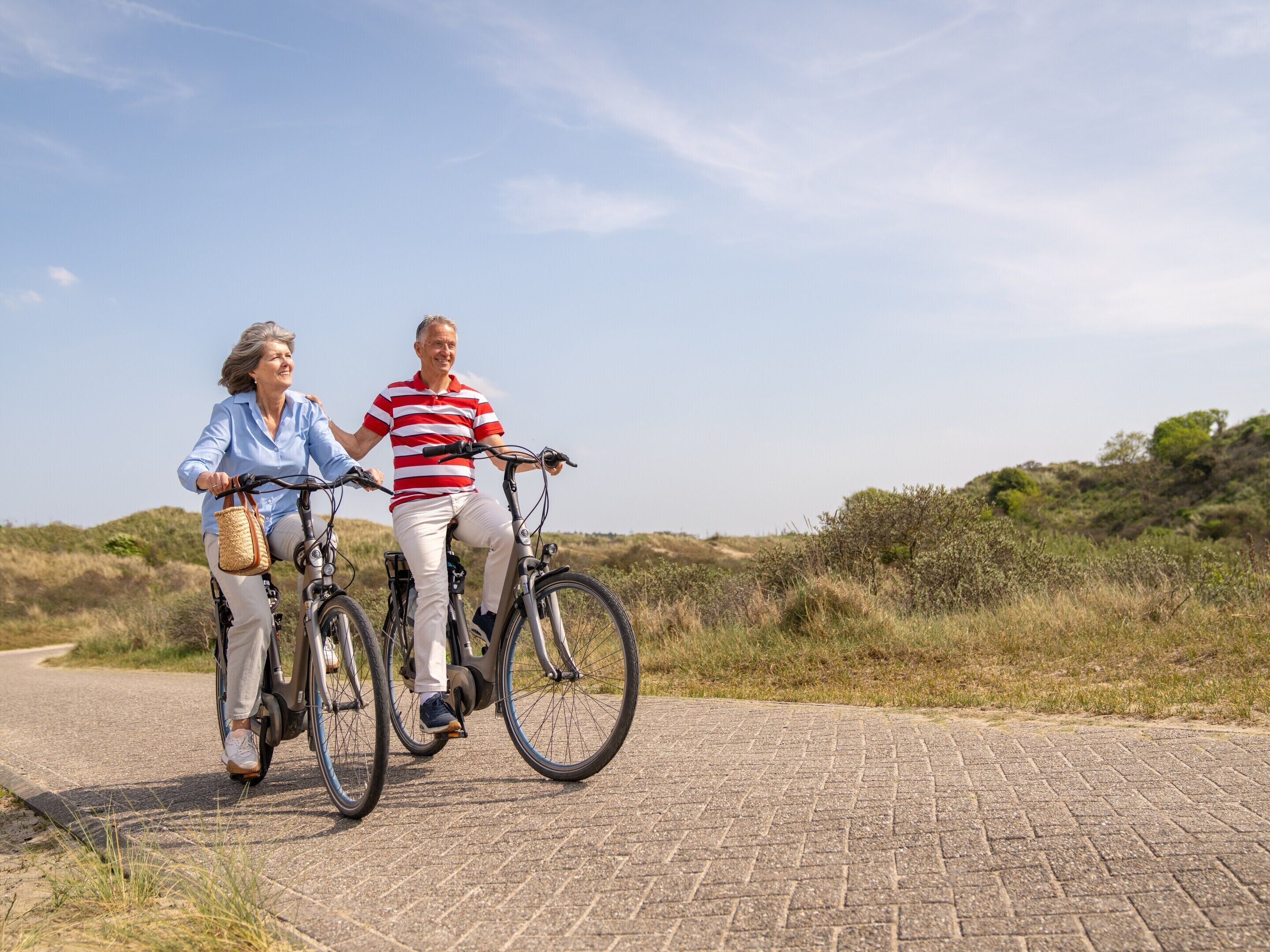 Two people riding bicycles on a paved path with a natural landscape in the background.