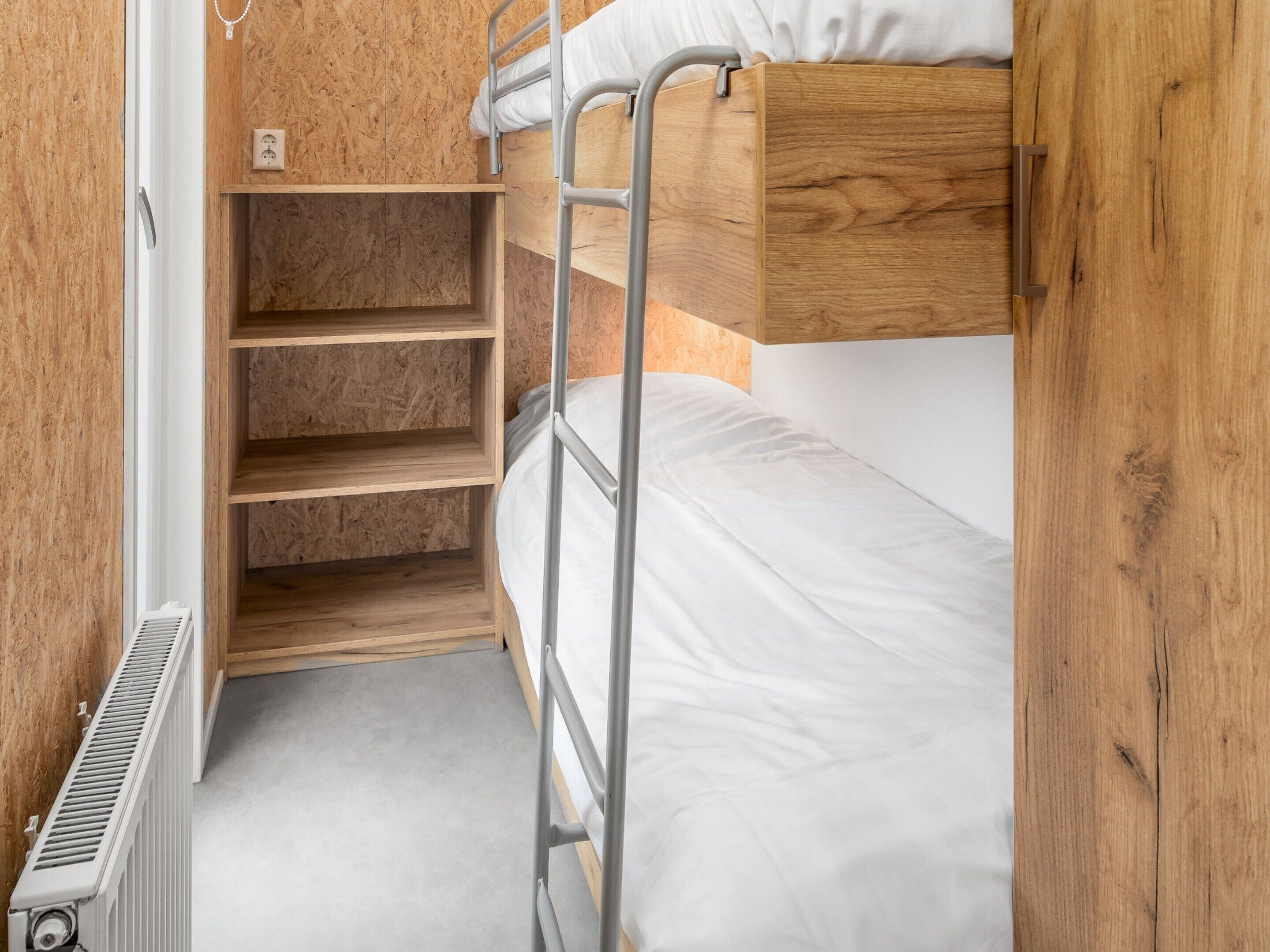 A bunk bed with a wooden headboard and a metal ladder. There is a white mattress and a shelf below the bed.