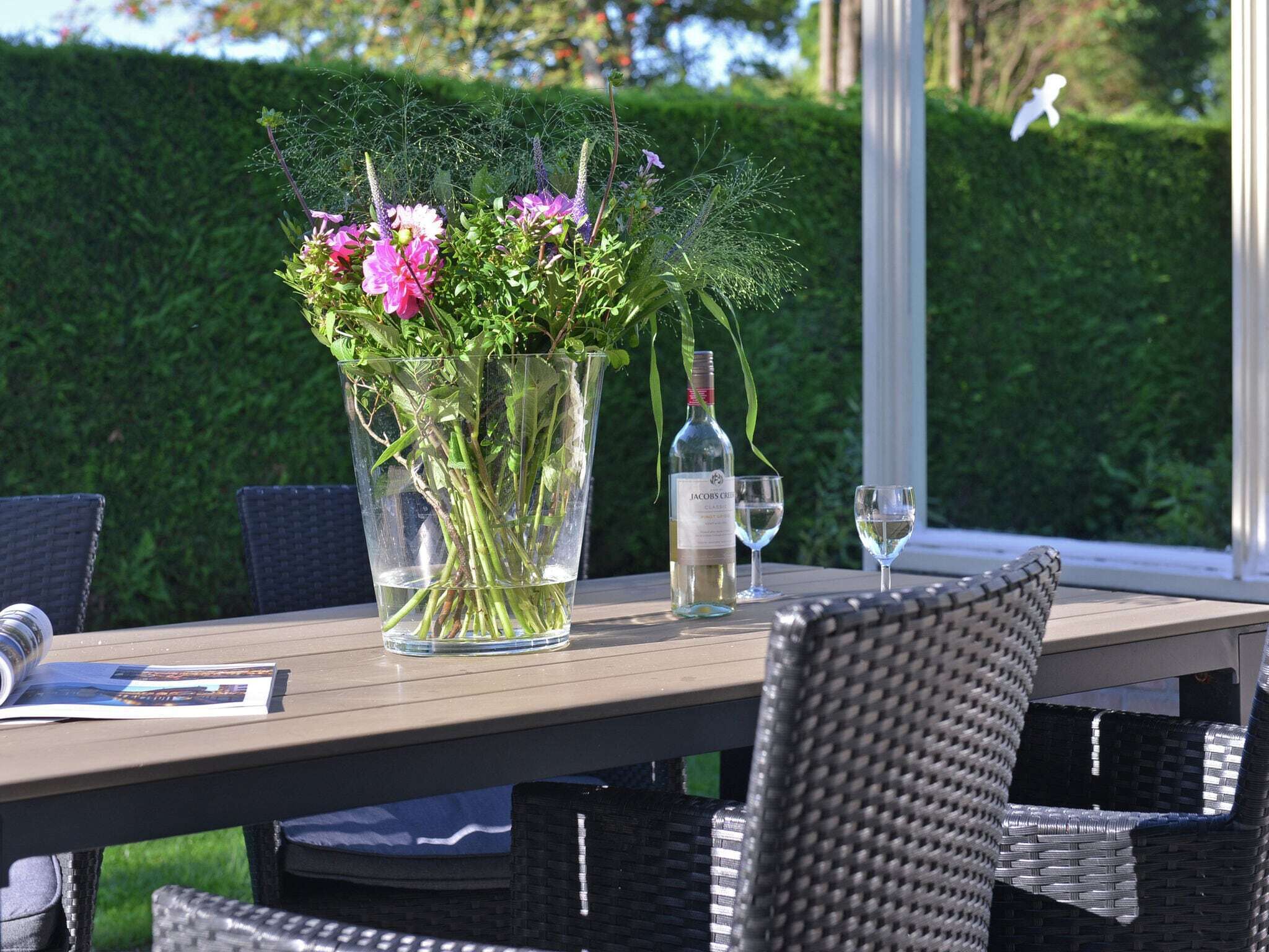 Ett bord med en vas av glas fylld med blommor, en vinflaska och två glas på ett träbord utomhus.