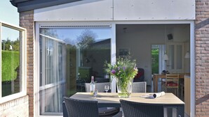 A patio with a table and chairs, a vase with flowers, and a sliding glass door leading to an indoor area. - Bungalow Near Veerse Meer & Beach (Kamperland)