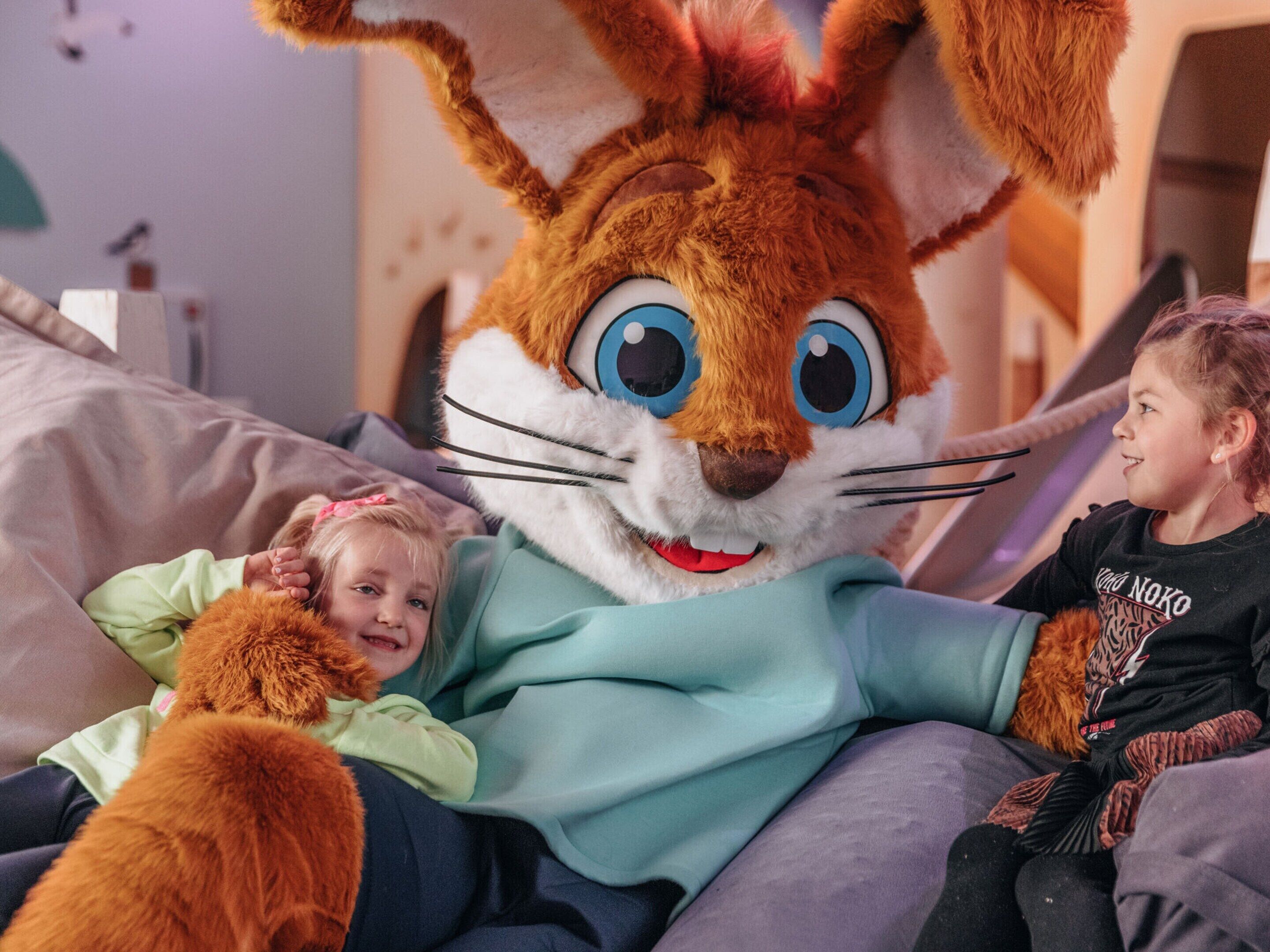Une personne vêtue d’un grand costume à fourrure, assise sur un lit avec deux enfants.