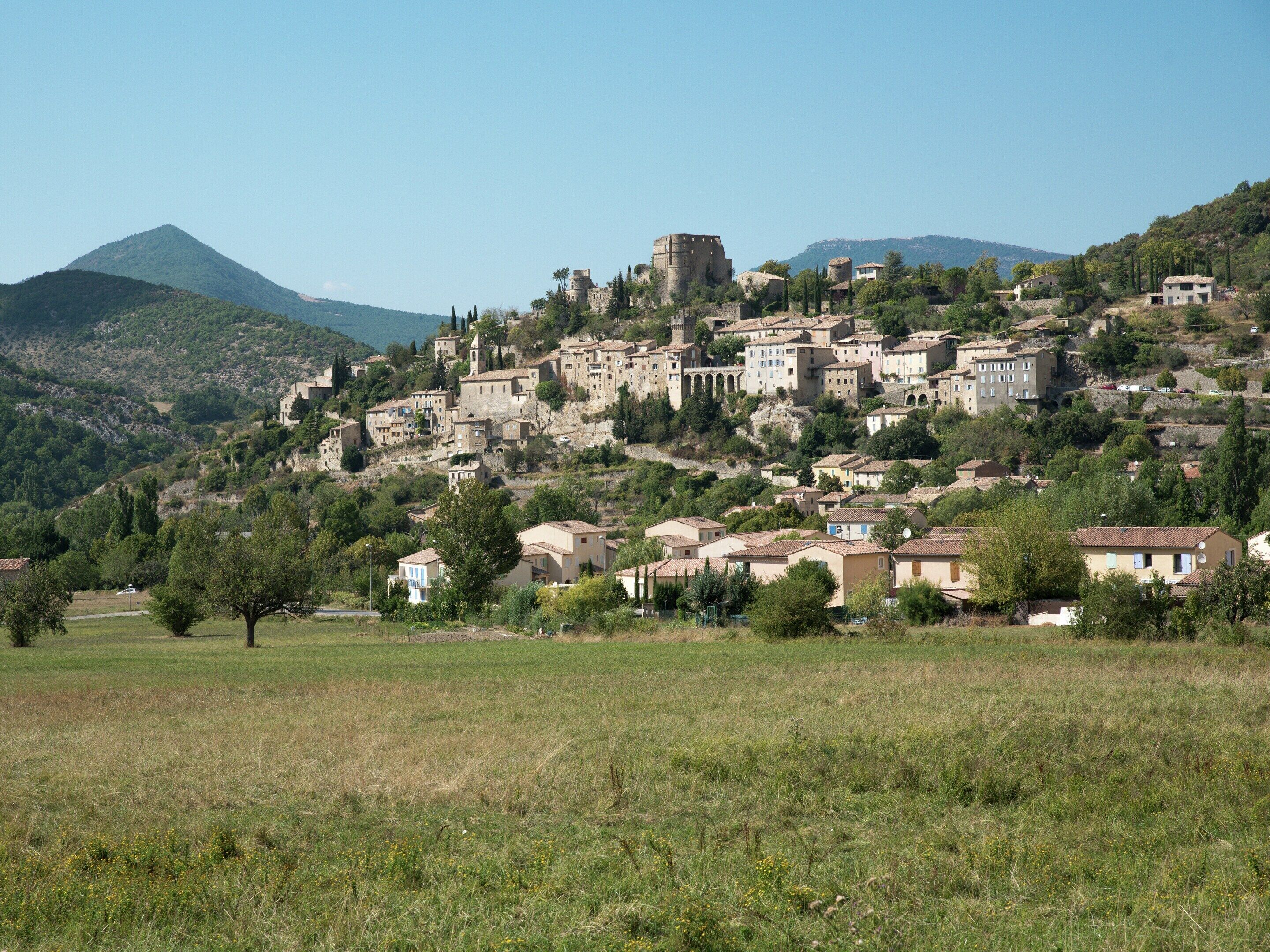 A hillside village with historic stone buildings, a prominent castle, and a clear view of the surrounding mountains.