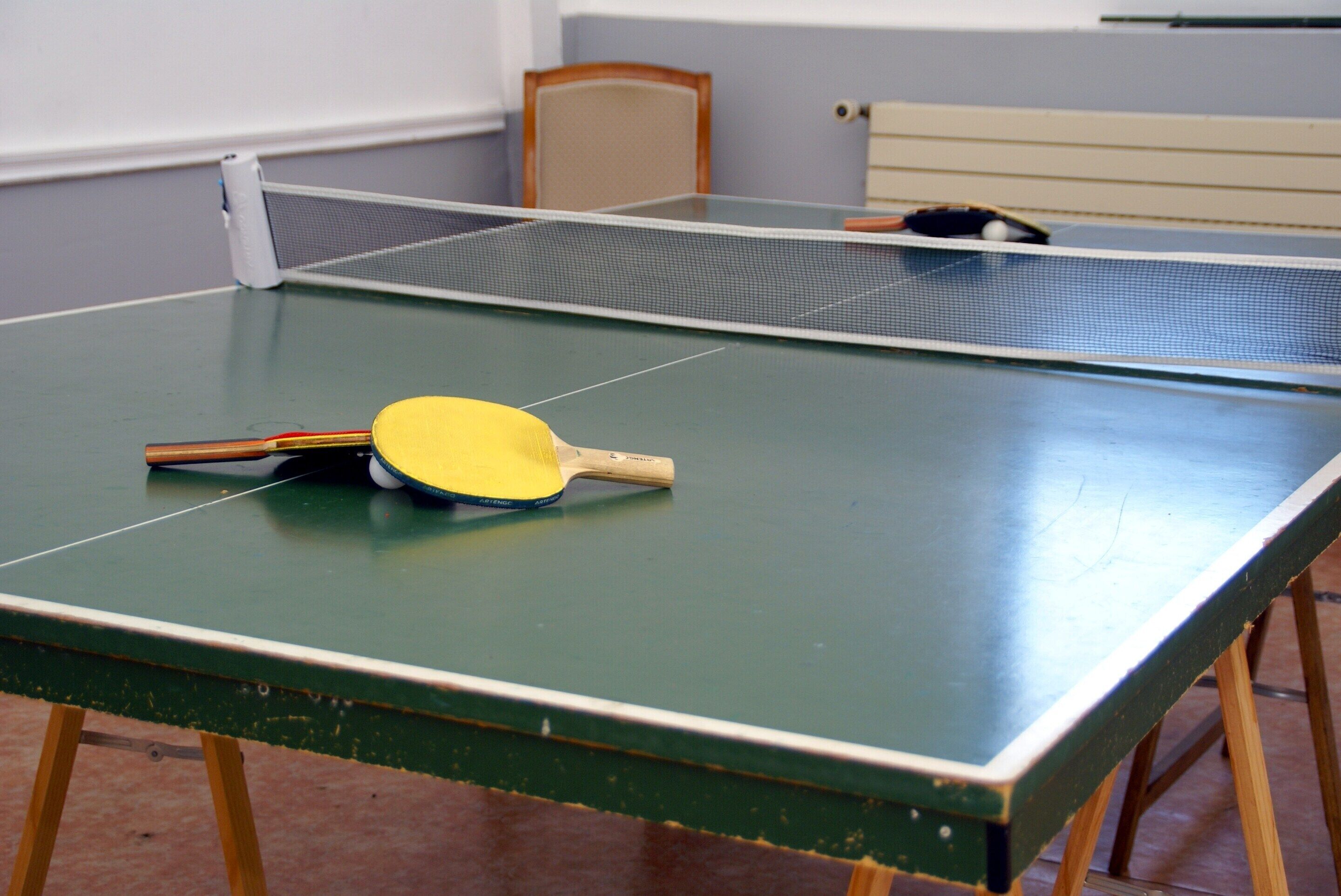 Egy pingpongasztal hálós ráccsal, egy szobában, ahol egy fa szék és a háttérben egy ágy látható.