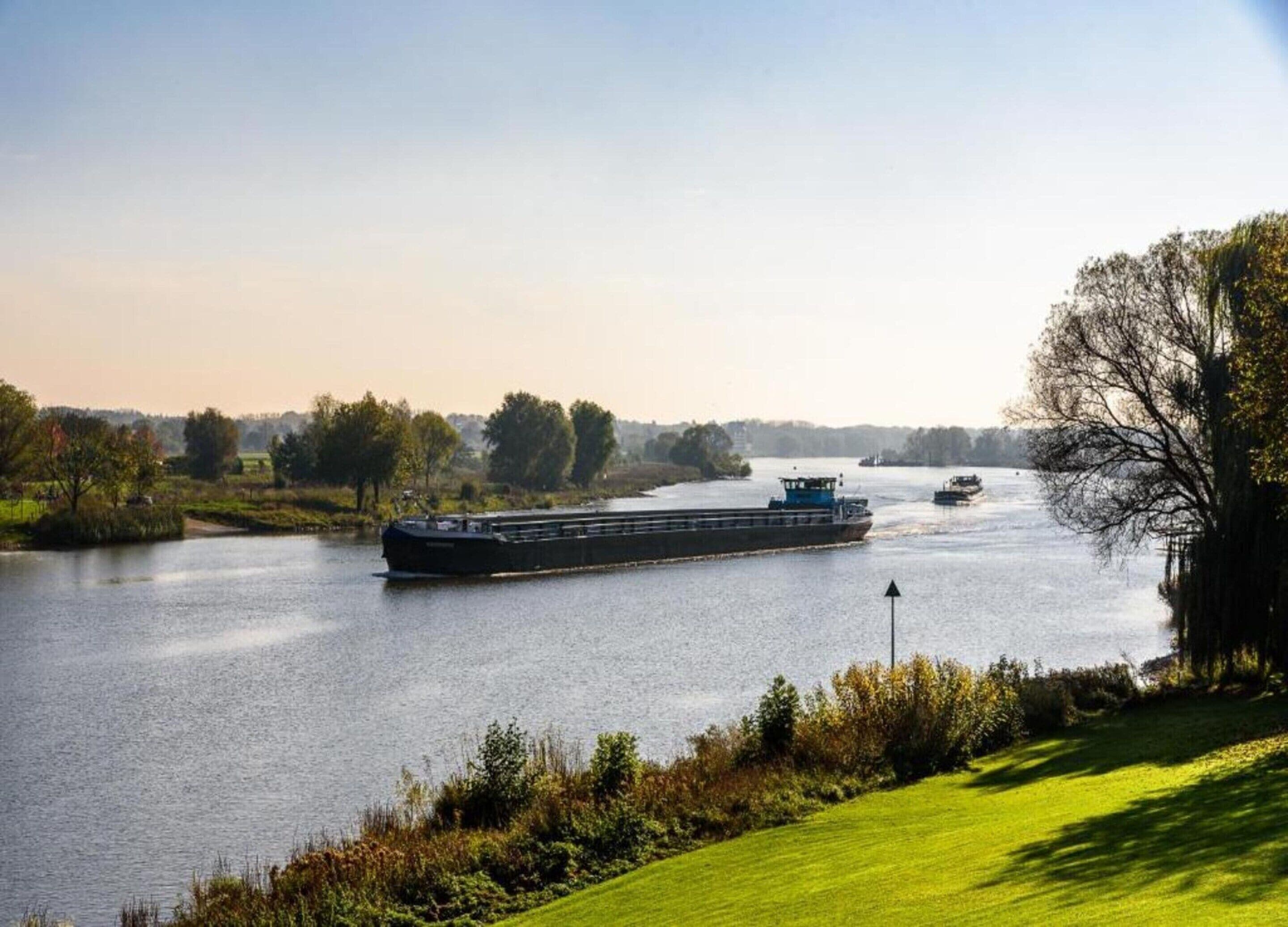 A river with a large cargo ship and smaller boats, surrounded by greenery and trees.