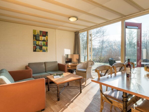 Ein gemütliches Wohnzimmer mit einer Couch, Sesseln, einem Couchtisch und einem Esstisch. Ein Fernseher, eine Lampe und ein Gemälde sind an der Wand zu sehen.