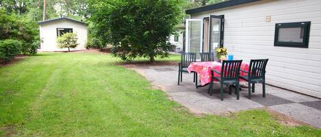 A small, single-story building with a patio area featuring a table and chairs, surrounded by a well-maintained lawn and trees.