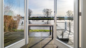 A view from a room with large glass doors onto a terrace with a bench, a metal railing, and a body of water in the background. - Holiday Home With Private Jetty on Water (Akkrum)